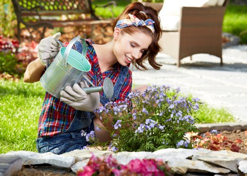 A young woman with light skin, wearing a blue and red plaid shirt, denim overalls, grey gardening gloves, and a colorful headband, is kneeling on a garden lawn during daylight hours. She is watering a small flowering shrub with light purple blooms using a metal watering can. The garden features a neatly maintained grassy area in the foreground, bordered by a stone edging, with flower beds containing various colorful plants and shrubs. In the background, there are outdoor furniture pieces, including a brown chair and a white bench, situated on a paved patio area, with a lush green lawn and mature trees providing natural shade. The scene depicts a well-kept outdoor space suitable for gardening and landscaping activities, consistent with professional gardening services available in Forest Gate, Epping Forest area, suggesting a typical suburban garden environment.
