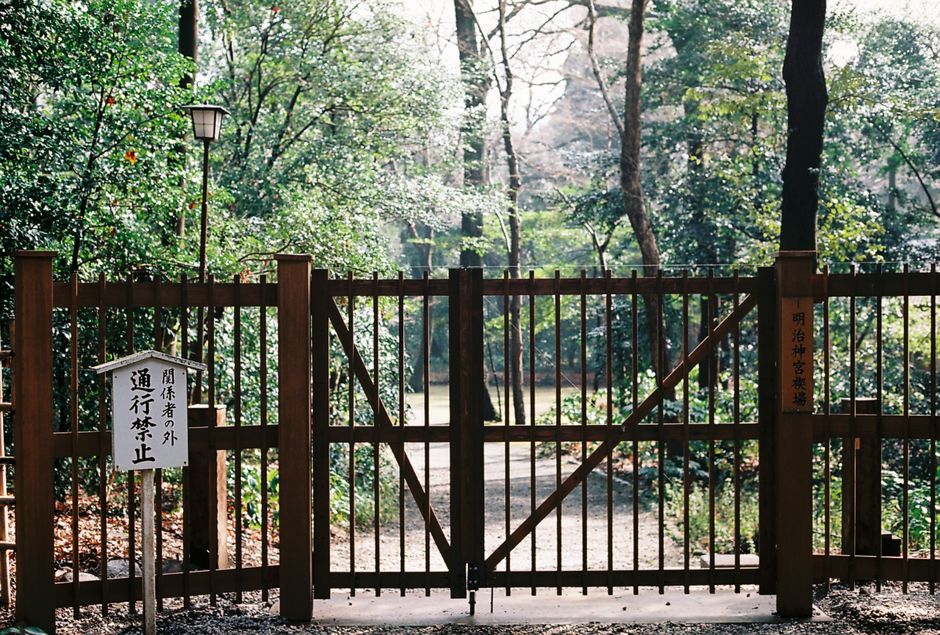 A black wrought iron gate with diamond-patterned lattice design, centered in a gravel pathway leading through a lush garden. The gate is flanked by two stone pillars and features a curved top arch. Be