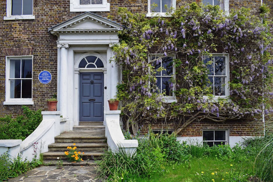 A charming front garden in Wanstead Flats featuring a classic brick townhouse with a white portico entrance and a blue painted door. The entrance is accessed via a short flight of stone steps with whi
